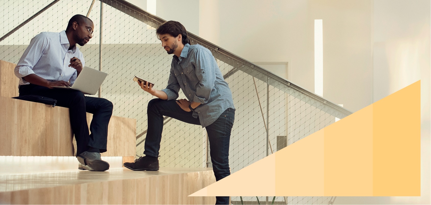 Two businessmen, one with a laptop and the other with a mobile phone, collaborate while sitting on a set of office auditorium steps.
